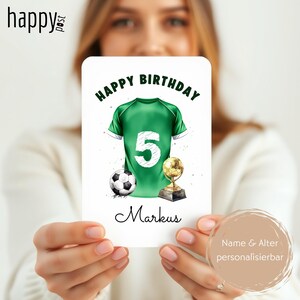 May include: A birthday card featuring a green soccer jersey with the number 5, a soccer ball, and a trophy. The words "Happy Birthday" are at the top, and the name "Markus" is at the bottom. The card is held by a person.