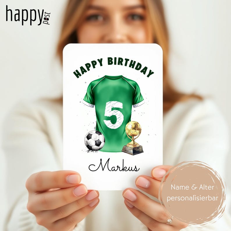 May include: A birthday card featuring a green soccer jersey with the number 5, a soccer ball, and a trophy. The words "Happy Birthday" are at the top, and the name "Markus" is at the bottom. The card is held by a person.