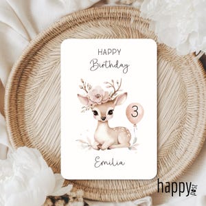 May include: A birthday card featuring a watercolor illustration of a fawn with a floral crown and a number 3 balloon. The card reads "Happy Birthday" and "Emilia". The card is on a woven tray.