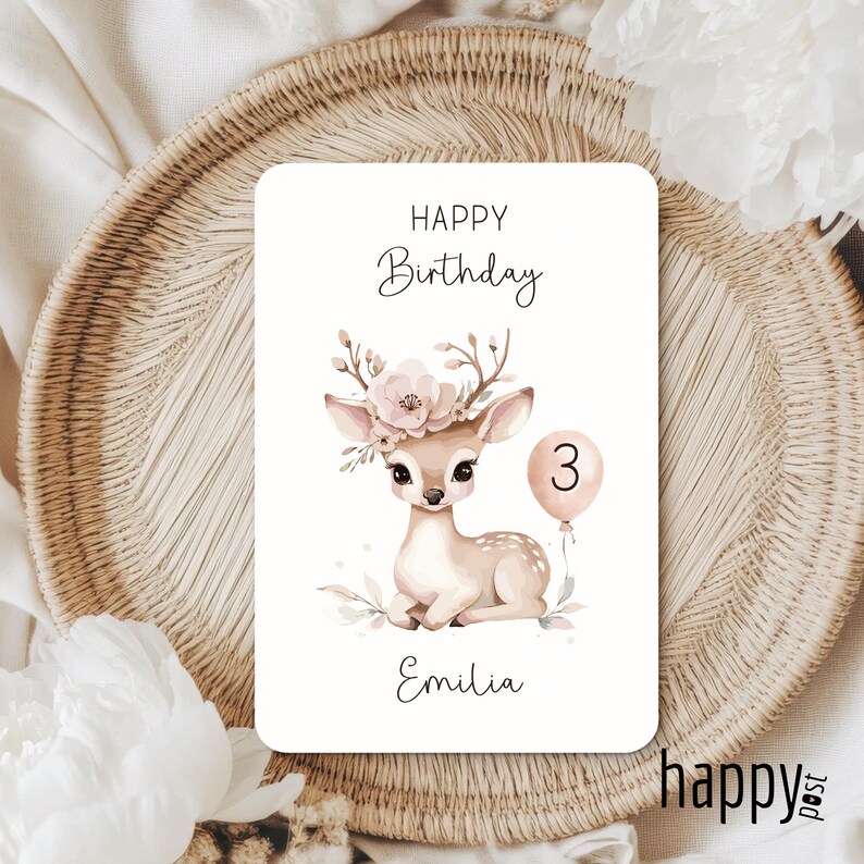 May include: A birthday card featuring a watercolor illustration of a fawn with a floral crown and a number 3 balloon. The card reads "Happy Birthday" and "Emilia". The card is on a woven tray.