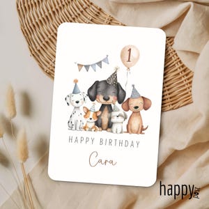 May include: A birthday card featuring five cartoon dogs wearing party hats. The card has the text "Happy Birthday Cara" and a balloon with the number "1". A banner with triangle flags is above the dogs. The card is on a neutral background.