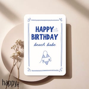 Pode incluir: Um cartão de felicitações retangular branco com texto azul que diz "HAPPY BIRTHDAY beach babe". O cartão apresenta uma borda azul e uma ilustração de biquíni. Está sobre um prato branco com flores secas.
