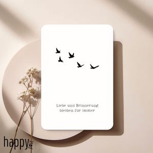 Puede incluir: Una tarjeta blanca con esquinas redondeadas presenta una bandada de cinco pájaros negros en vuelo. La tarjeta tiene el texto alemán "Liebe und Erinnerung bleiben für immer" impreso en una fuente sencilla. La tarjeta está sobre un plato beige con una ramita de flores blancas.