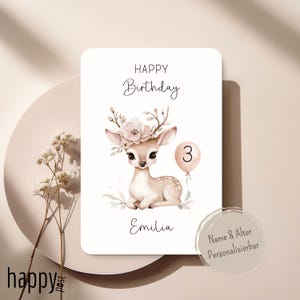 May include: A birthday card featuring a watercolor illustration of a fawn with a floral crown and a balloon with the number 3. The card has the words "Happy Birthday" and the name "Emilia" written on it. The card is on a light beige background.