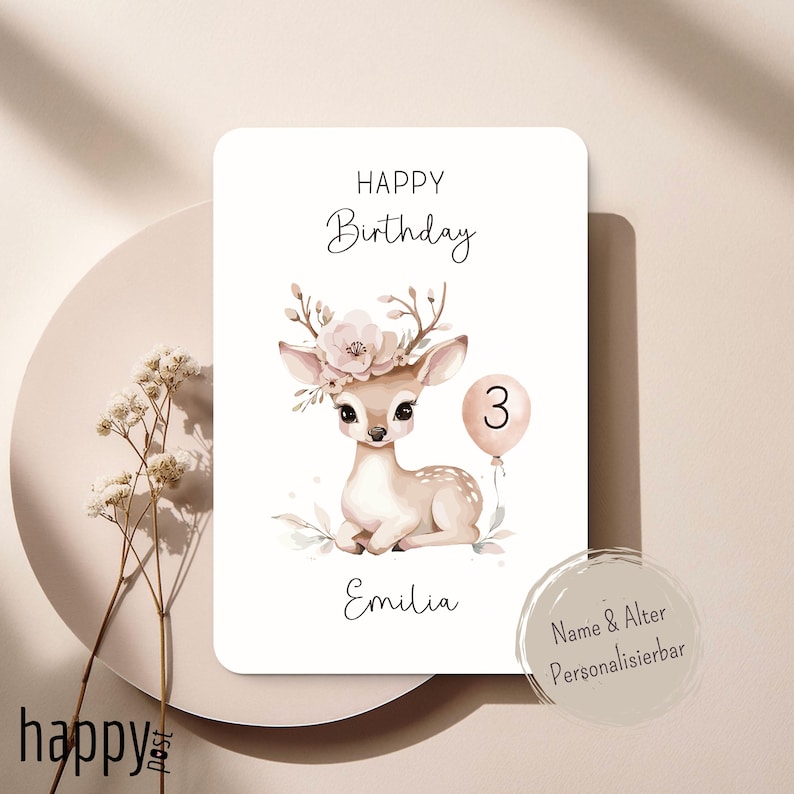 May include: A birthday card featuring a watercolor illustration of a fawn with a floral crown and a balloon with the number 3. The card has the words "Happy Birthday" and the name "Emilia" written on it. The card is on a light beige background.