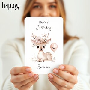 May include: A birthday card featuring a fawn with a floral crown and a number 3 balloon. The card reads "Happy Birthday" and "Emilia" in elegant script. The fawn is light brown with white spots.
