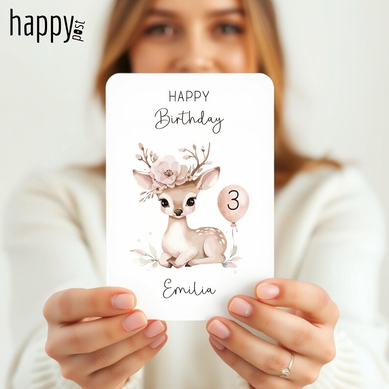 May include: A birthday card featuring a fawn with a floral crown and a number 3 balloon. The card reads "Happy Birthday" and "Emilia" in elegant script. The fawn is light brown with white spots.