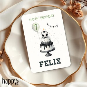 May include: A birthday greeting card with a soccer ball cake design. The card reads "Happy Birthday" and "Felix". A balloon with the number 7 is also featured. The card is on a white plate with a gold rim.