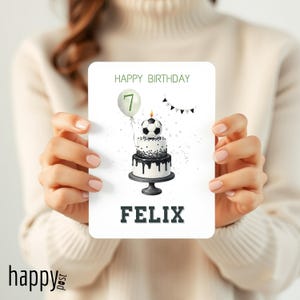 May include: A birthday card featuring a soccer ball cake with a lit candle and a balloon with the number 7. The card reads "HAPPY BIRTHDAY" and "FELIX" in green and black text. The card is held by a person.