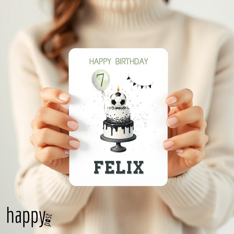 May include: A birthday card featuring a soccer ball cake with a lit candle and a balloon with the number 7. The card reads "HAPPY BIRTHDAY" and "FELIX" in green and black text. The card is held by a person.