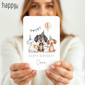 May include: A birthday card featuring illustrations of five dogs wearing party hats. The card has the text "Happy Birthday Cara" and a balloon with the number "1". A banner with gray and blue triangles is also present.