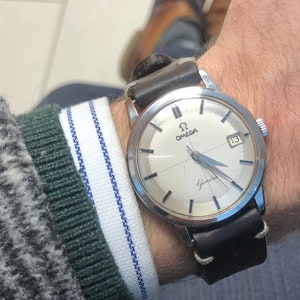 May include: A silver wristwatch with a white face and black leather strap. The watch face has black Roman numeral hour markers, black hands, and a date window at the 3 o'clock position. The watch face also has the text "Omega" and "Geneva" printed on it.