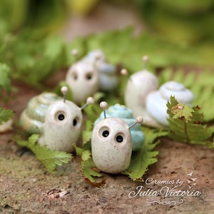 May include: A collection of small ceramic snail figurines with speckled off-white bodies, dark eyes, and thin antennae. The shells are glazed in shades of light blue and green. The text "Ceramics by Julia Victoria" is printed in the bottom right corner.