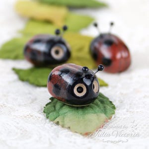 Può includere: Tre piccole statuette di coccinelle in ceramica con corpi lucidi neri e rossi, macchie nere e due sottili antenne. Ogni coccinella presenta grandi occhi rotondi chiari ed è posizionata su una foglia di tessuto verde. Testo: Ceramics by Julia Victoria.