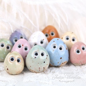 May include: A collection of small, egg-shaped ceramic figurines with glossy finishes, each featuring two prominent black and white eyes. The figurines come in various pastel colors, including blue, pink, green, and cream, with some speckled details.