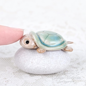Peut inclure: Une petite figurine de tortue en céramique avec une carapace brillante bleu clair et verte et une tête beige, posée sur une pierre grise lisse et arrondie. La tortue a un œil noir et est placée près d'un doigt. Texte : Ceramics by Julia Victoria.
