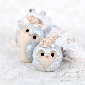 May include: Two ceramic owl figurines with light blue speckled bodies and cream-colored faces. Each owl features a small, tan-colored seashell attached to the top of its head. The owls have black eyes and a small, pointed, gold-colored beak.