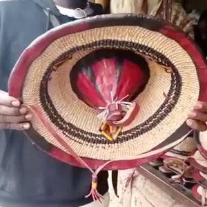 以下が含まれることがあります： 赤、黒、白の縁取りが施された、編まれた麦わら帽子。帽子には革のストラップとタッセルが付いています。