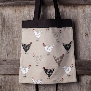 Könnte beinhalten: Eine beigefarbene Einkaufstasche mit schwarzen Henkeln und einem sich wiederholenden Muster aus schwarzen und weißen Hühnern mit roten Kämmen. Die Tasche hängt an einer Holzwand.