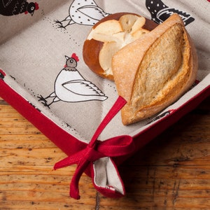 May include: A red fabric bread basket with a white and black chicken pattern. The basket is filled with two rolls, one pretzel shaped and one round.