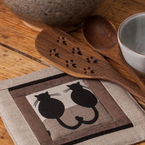 Pode incluir: Um porta-panelas de linho marrom com um design de gato preto. O porta-panelas tem um laço para pendurar. Duas colheres de madeira com desenhos de pegadas de patas estão no fundo.