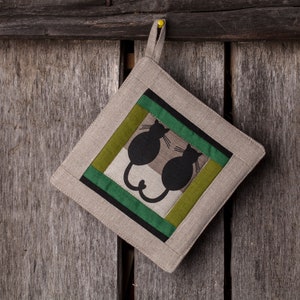 Può includere: Un sottopentola quadrato con un design a patchwork verde, marrone e beige. Il sottopentola presenta una silhouette nera di due gatti seduti uno di fronte all'altro.