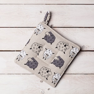 以下が含まれることがあります： 黒と白の羊の模様がついたベージュの鍋つかみ。鍋つかみには吊り下げ用のループが付いています。