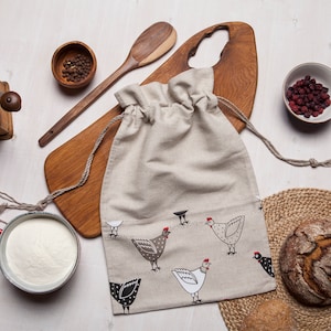 Puede incluir: Una bolsa de cordón de lino beige con ilustraciones de pollos en blanco y negro. La bolsa está sobre una tabla de cortar de madera con una cuchara de madera y un tazón de granos de pimienta. También hay un tazón de arándanos secos y una hogaza de pan sobre un mantel tejido.