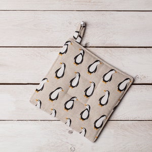 Puede incluir: Un agarrador de olla beige con un patrón de pingüinos. El agarrador de olla tiene un lazo para colgar.