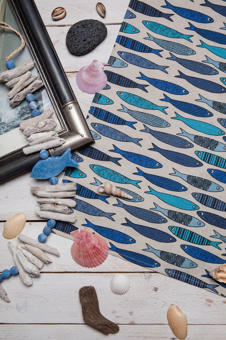 May include: A white background with a blue and white fabric with a pattern of blue fish. There are seashells, driftwood, and beads scattered around the fabric.