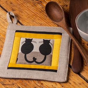 Può includere: Un sottopentola in lino beige con un bordo giallo e nero. Al centro, due silhouette di gatti neri si guardano con le code intrecciate.