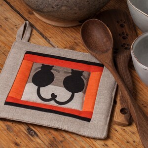 May include: A beige linen pot holder with a black and orange frame and two black cats inside. The cats are facing each other and their tails are intertwined.