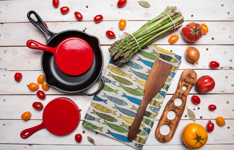 May include: A set of two red cast iron skillets with black handles, a wooden spatula, a wooden olive oil dispenser, a bunch of asparagus, and various vegetables on a white wooden surface.
