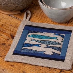 May include: A square, beige linen pot holder with a blue and black geometric pattern featuring a design of blue fish. The pot holder has a loop for hanging.