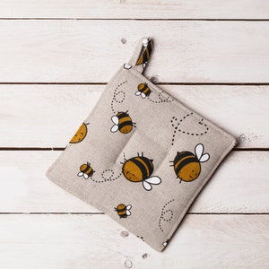 Può includere: Un sottopentola in lino beige con un motivo a forma di ape. Le api sono gialle e nere e volano in un motivo a linea punteggiata.