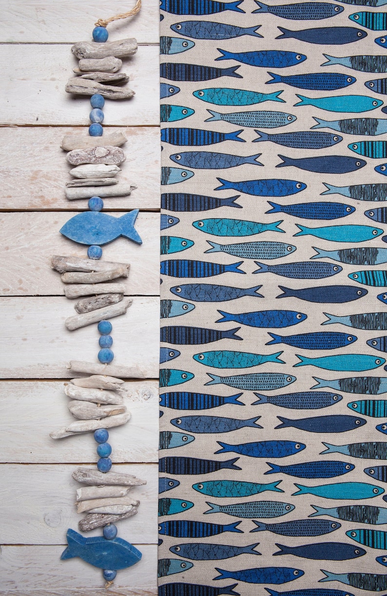 May include: A blue and white wooden garland with driftwood and blue fish shapes. The garland is hanging on a white wooden background. The background is a fabric with a pattern of blue fish.