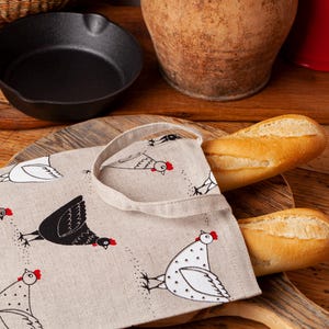 Pode incluir: Uma bolsa de lona bege com estampa de frango preto e branco. A bolsa está cheia de duas baguetes longas.