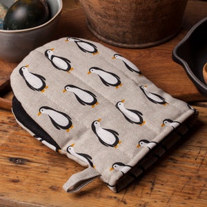 Puede incluir: Un guante de horno beige con ribete negro y un patrón de pingüinos negros y blancos. El guante tiene un asa de bucle.