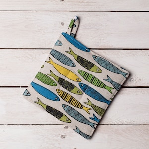 Puede incluir: Un agarrador de olla blanco con un patrón de peces azules, verdes y amarillos. El agarrador de olla tiene un lazo para colgar.