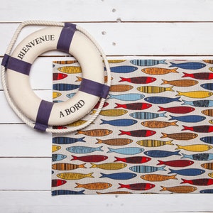 Op de afbeelding: Een wit en blauwe reddingsboei met de tekst "Bienvenue a Bord" ernaast een beige placemat met een patroon van kleurrijke vissen.