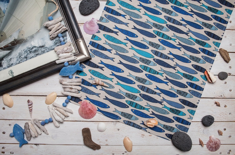 May include: A blue and white kitchen towel with a pattern of fish. The towel is lying on a white wooden surface with driftwood, seashells, and stones.