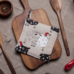 以下が含まれることがあります： 赤いサンタの帽子をかぶった茶色と黒の羊の鍋つかみ2個セット。鍋つかみは木製のまな板の上に置かれ、背景には木製のさじと胡椒の入ったボウルがある。