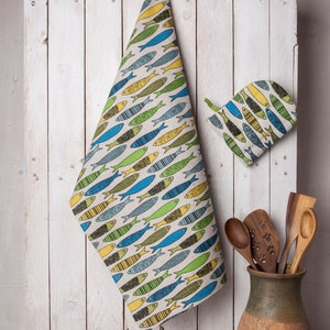 May include: A set of kitchen towels with a colorful fish pattern. The towels are hanging on a white wooden wall. There is a matching pot holder and wooden spoons in a brown ceramic pot.