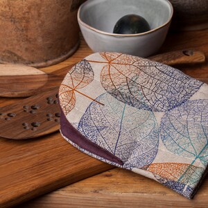 Puede incluir: Un guante de horno marrón y beige con un patrón de hojas. El guante tiene un borde marrón oscuro y está hecho de una tela suave y texturizada.