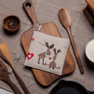 Op de afbeelding: Een houten snijplank met een bruine linnen ovenwant met daarop twee rendieren met rode harten. De ovenwant ligt op de snijplank. Er liggen andere keukengerei op een bruine linnen ondergrond.
