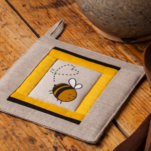 May include: A square beige linen pot holder with a yellow and black border. The centre features a cartoon bee with black and yellow stripes and a heart-shaped trail behind it.