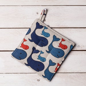 Puede incluir: Un juego de dos agarraderas de olla de color azul y blanco con un patrón de ballenas y un detalle de ballena roja. Las agarraderas tienen un lazo para colgar.