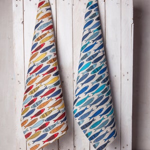 May include: Two kitchen towels hanging on a white wooden wall. The towels are made of linen and feature a pattern of colorful fish. One towel is yellow, orange, red, and blue. The other towel is blue and teal.
