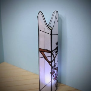 May include: A white stained glass lamp with a floral design. The lamp is shaped like a tall, narrow cylinder with a pointed top. The floral design features brown branches and pink flowers.
