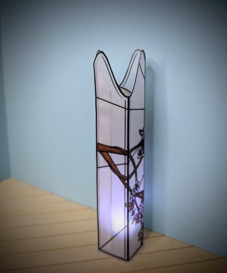 May include: A white stained glass lamp with a floral design. The lamp is shaped like a tall, narrow cylinder with a pointed top. The floral design features brown branches and pink flowers.